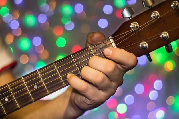 Uomo che suona la chitarra acustica. Le dita del primo piano sul collo della chitarra contro natale hanno offuscato le luci del bokeh su fondo.