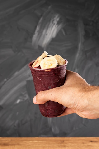 Uomo che tiene il frullato di gelato brasiliano congelato AÃ§ai Berry in tazza di plastica con banane. Su una scrivania in legno e uno sfondo estivo grigio con frutta. Vista frontale per menu e social media