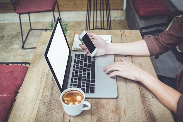 uomo che tiene smartphone sul tavolo con computer portatile e caffè