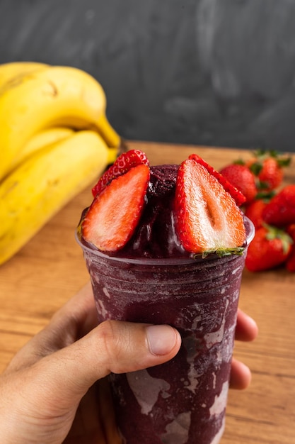 Uomo che tiene un frullato di gelato brasiliano AÃ§ai Berry congelato in tazza di plastica con fragole. Su una scrivania in legno e uno sfondo estivo grigio con frutta. Vista frontale per menu e social media