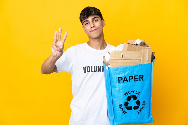 Uomo che tiene un sacchetto di riciclaggio pieno di carta da riciclare sopra fondo giallo isolato felice e contando tre con le dita
