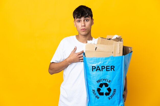 Uomo che tiene una borsa per il riciclaggio piena di carta da riciclare su giallo sorpreso e scioccato mentre guarda a destra