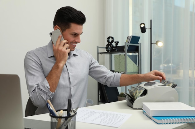 Uomo che utilizza il contatore di banconote mentre parla al telefono al tavolo bianco all'interno