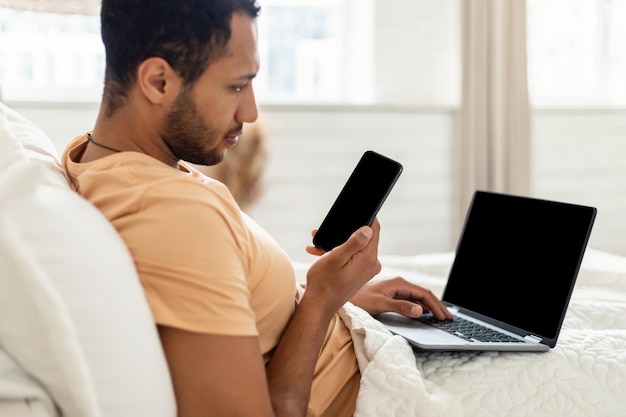 Uomo che utilizza telefono e laptop seduto a letto a casa