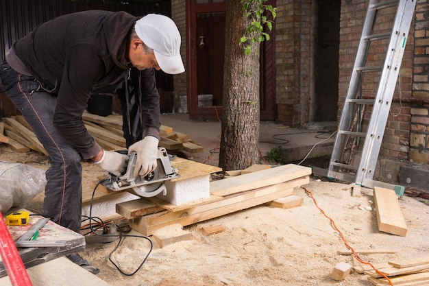 Uomo che utilizza una sega portatile per tagliare assi di legno per la costruzione di case lasciando dietro di sé pile di polvere di sega sul pavimento