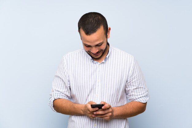 Uomo colombiano sopra isolato muro blu inviando un messaggio con il cellulare