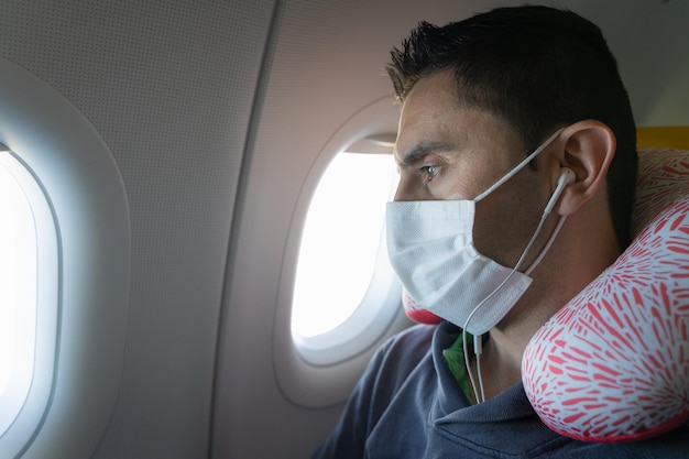 Uomo con auricolari con maschera facciale e cuscino per il collo che guarda attraverso la finestra dell'aereo Misure sanitarie dell'aeroplano