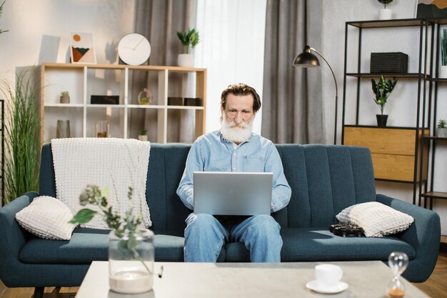 Uomo con barba grigia maschio maturo in abiti casual seduto sul divano morbido blu con il computer portatile