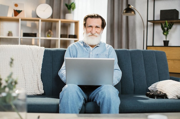 Uomo con barba grigia maschio maturo in abiti casual seduto sul divano morbido blu con il computer portatile