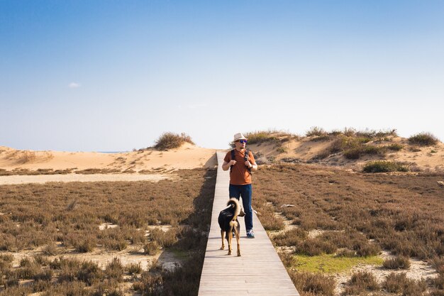 Uomo con cane che cammina sul sentiero di legno sulla spiaggia e guarda in lontananza l'oceano.