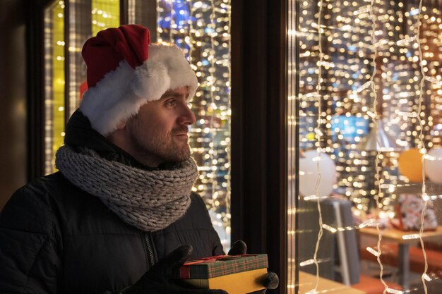 Uomo con cappello di Babbo Natale che guarda e sogna nella vetrina illuminata del negozio natalizio concetto di vendita