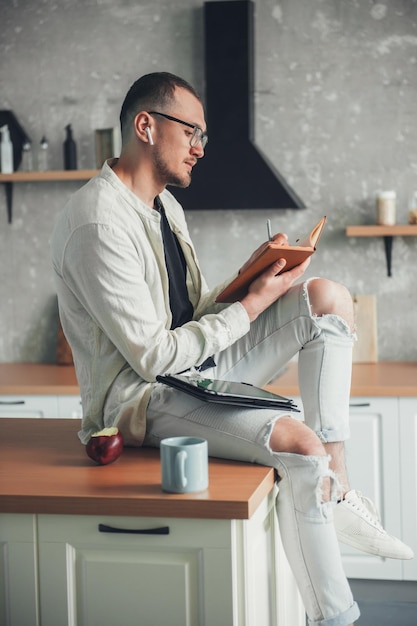 Uomo con gli occhiali seduto in cucina a prendere appunti durante una videoconferenza con i colleghi Lavoro a distanza da casa in un appartamento moderno Formazione online