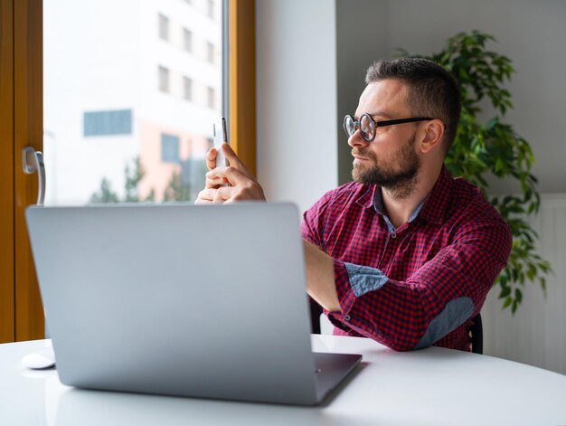 Uomo con gli occhiali seduto in ufficio a casa e lavorando su un laptop e utilizzando lo smartphone