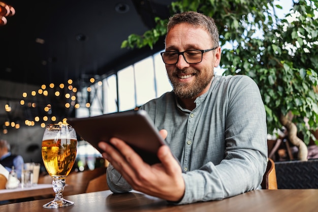 Uomo con gli occhiali seduto in un bar dopo il lavoro e utilizzando il tablet per appendere sui social media.