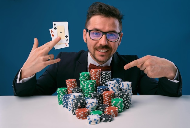 Uomo con gli occhiali vestito nero seduto al tavolo bianco con pile di fiches in possesso di due carte da gioco pos...