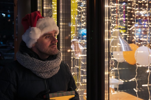 Uomo con il cappello di Babbo Natale con una scatola di regali vicino alla finestra illuminata del caffè