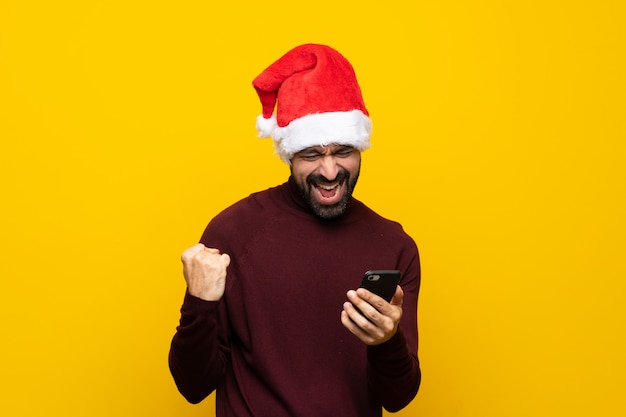 Uomo con il cappello di natale sopra fondo giallo isolato con il telefono nella posizione di vittoria