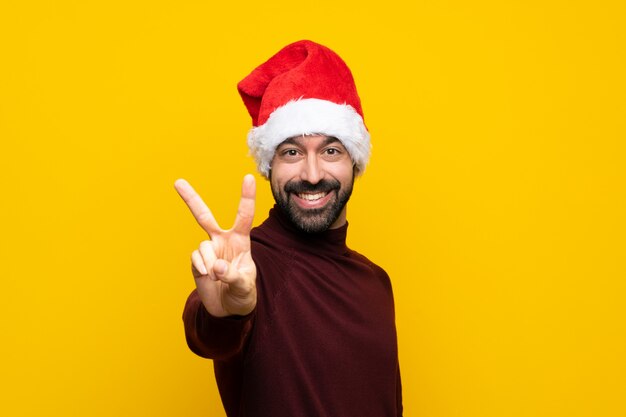 Uomo con il cappello di natale sopra la parete gialla isolata che sorride e che mostra il segno di vittoria