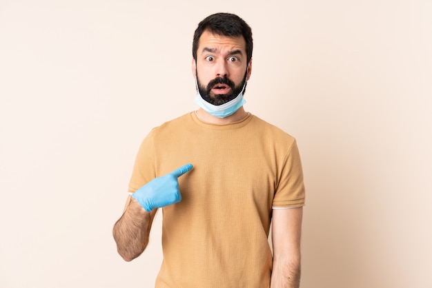 Uomo con la barba che protegge dal coronavirus con una maschera e guanti sopra la parete isolata che punta a se stessi