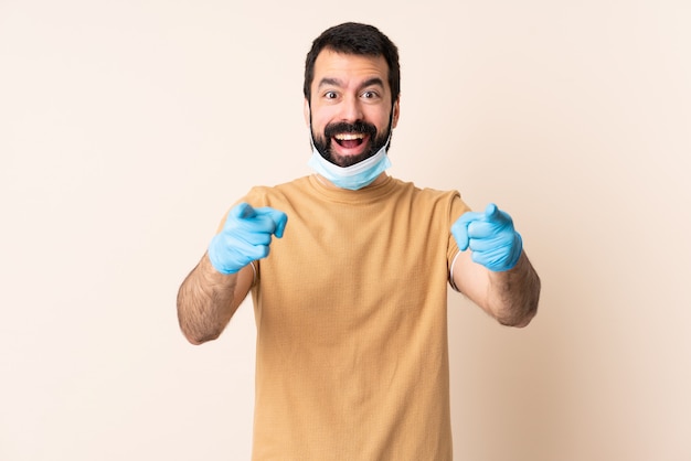Uomo con la barba che protegge dal coronavirus con una maschera e guanti sopra la parete isolata sorpresa e che indica parte anteriore
