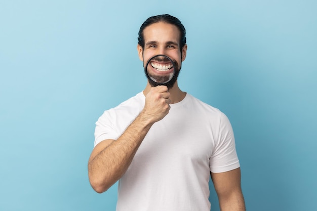 Uomo con la barba che tiene la lente d'ingrandimento sui denti guardando la fotocamera con un'espressione felice