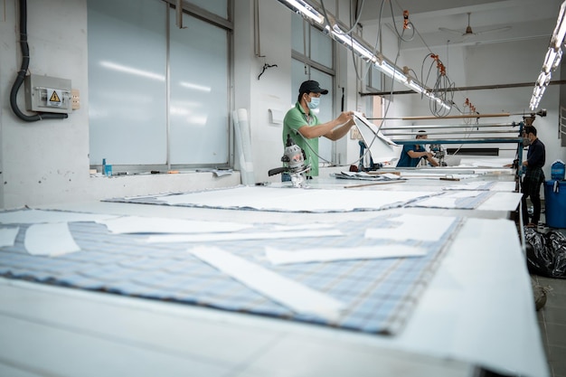 Uomo con macchina da taglio e dispositivi di protezione individuale presso il luogo di lavoro industriale di abbigliamento Taglierina per tessuti in una fabbrica di abbigliamento tessile asiatico