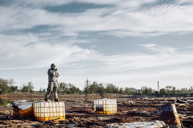 Uomo con maschera antigas e abiti militari verdi esplora i barili dopo il disastro chimico