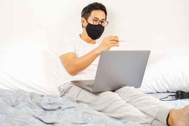 Uomo con maschera protettiva nel letto guardando termometro coronavirus in quarantena.