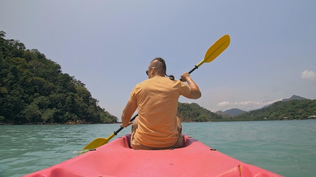 Uomo con occhiali da sole righe rosa canoa di plastica lungo il mare contro verdi isole collinari con giungle selvagge