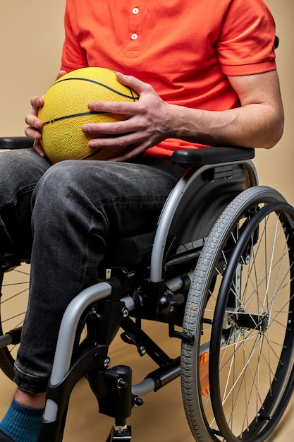 Uomo con palla da basket seduto su una sedia a rotelle contro il muro di colore, sport per disabili. foto in primo piano