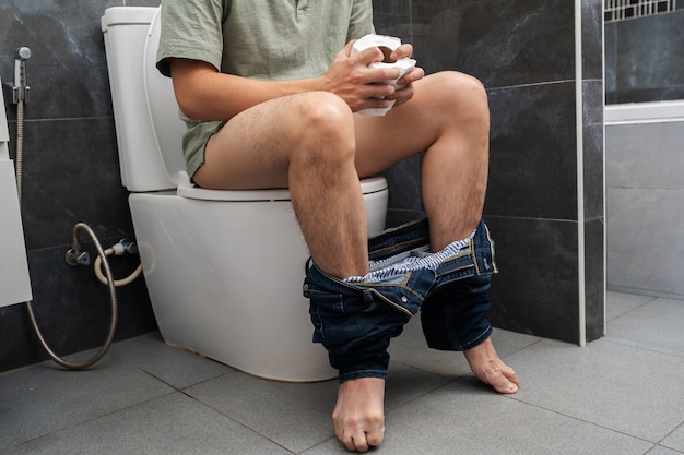 Uomo con problemi di stomaco seduto nella tazza del water e con in mano un rotolo di fazzoletti in bagno