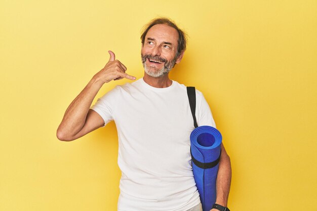 Uomo con tappetino yoga in studio giallo che mostra un gesto di chiamata sul cellulare con le dita