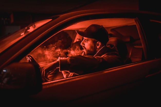 Uomo con un berretto alla guida di un'auto che fuma di notte in un garage illuminato da una luce rossa