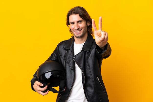 Uomo con un casco da motociclista isolato su sfondo giallo che sorride e che mostra il segno di vittoria