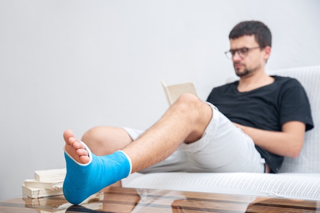 Uomo con una gamba rotta in stecca blu per il trattamento delle lesioni da distorsione alla caviglia leggendo libri presso la riabilitazione domestica.