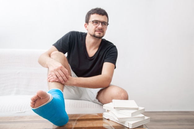 Uomo con una gamba rotta in stecca blu per il trattamento delle lesioni da distorsione alla caviglia leggendo libri presso la riabilitazione domestica.