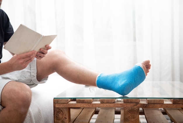 Uomo con una gamba rotta in stecca blu per il trattamento delle lesioni da distorsione alla caviglia leggendo libri presso la riabilitazione domestica.