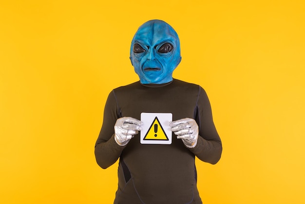 Uomo con una maschera di un alieno con una testa blu che tiene un cartello con il segno di pericolo e un punto esclamativo Concetto di bizzarro extraterrestre divertente informativo strano e strano