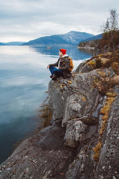 Uomo con uno zaino giallo che indossa un cappello rosso seduto sulla riva sullo sfondo della montagna e del lago