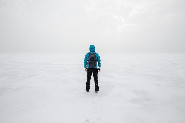 Uomo con zaino in piedi da solo sul ghiaccio del lago e guardando in avanti. Giornata invernale fredda e nuvolosa.