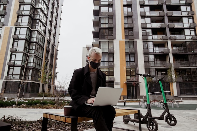 Uomo concentrato libero professionista in maschera facciale nera che lavora fuori nel parco e utilizza il suo laptop moderno. Si siede sulla panchina e scrive un programma. Due scooter elettrici vicino al banco. Blocchi di appartamenti.