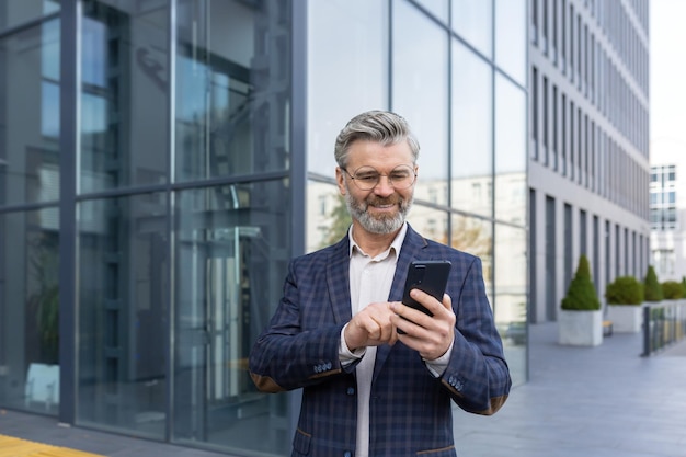 Uomo d'affari adulto maturo fuori dall'edificio per uffici che cammina e usa il capo dello smartphone digitando il messaggio