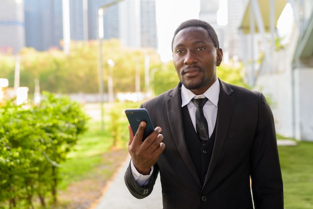 Uomo d'affari africano barbuto bello che pensa mentre usando telefono al parco