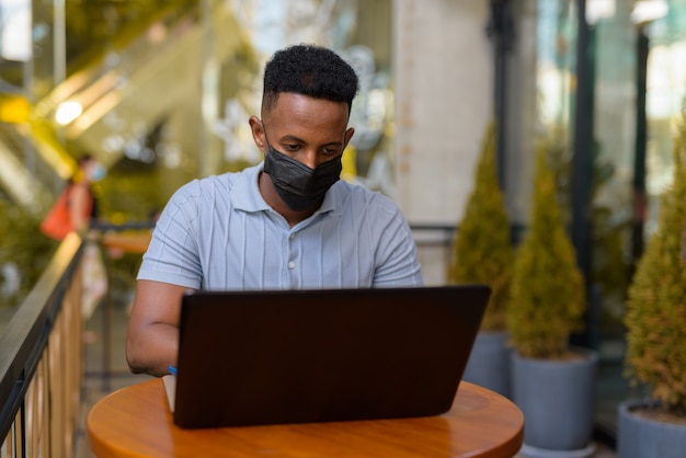Uomo d'affari africano che indossa maschera facciale e distanza sociale mentre è seduto al bar utilizzando il computer portatile