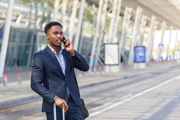 Uomo d'affari africano che viaggia vicino all'aeroporto con la valigia che chiama taxi utilizzando l'app mobile