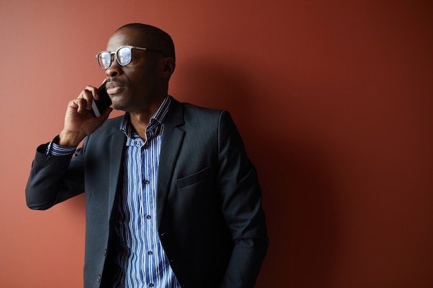 Uomo d'affari africano in occhiali da vista e in tuta parlando al telefono cellulare mentre in piedi contro lo sfondo marrone