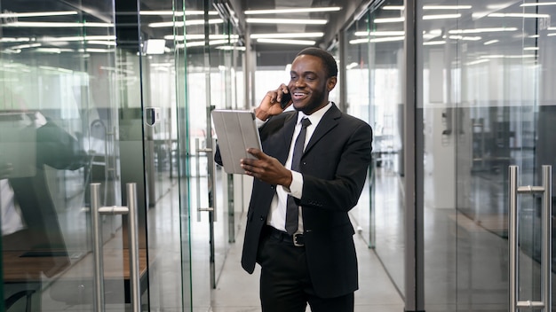 Uomo d'affari afroamericano che parla sullo smartphone mentre camminando nell'edificio per uffici corporativo e controllando i messaggi di posta elettronica online al pc della compressa. Concetto di uomo d'affari di successo.