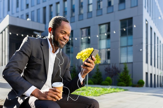 Uomo d'affari afroamericano che usa il telefono seduto sulla panchina fuori dall'edificio per uffici che guarda il capo