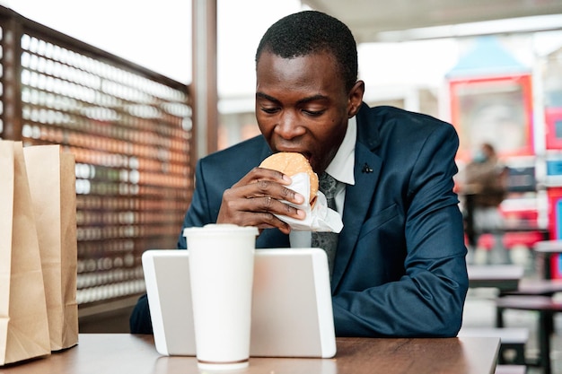 Uomo d'affari afroamericano in giacca e cravatta seduto nel ristorante a mangiare hamburger e utilizzando tablet