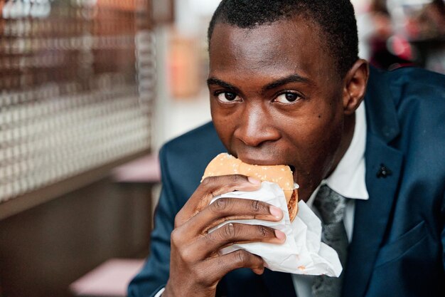 Uomo d'affari afroamericano mordere gustoso hamburger fresco e guardando la fotocamera